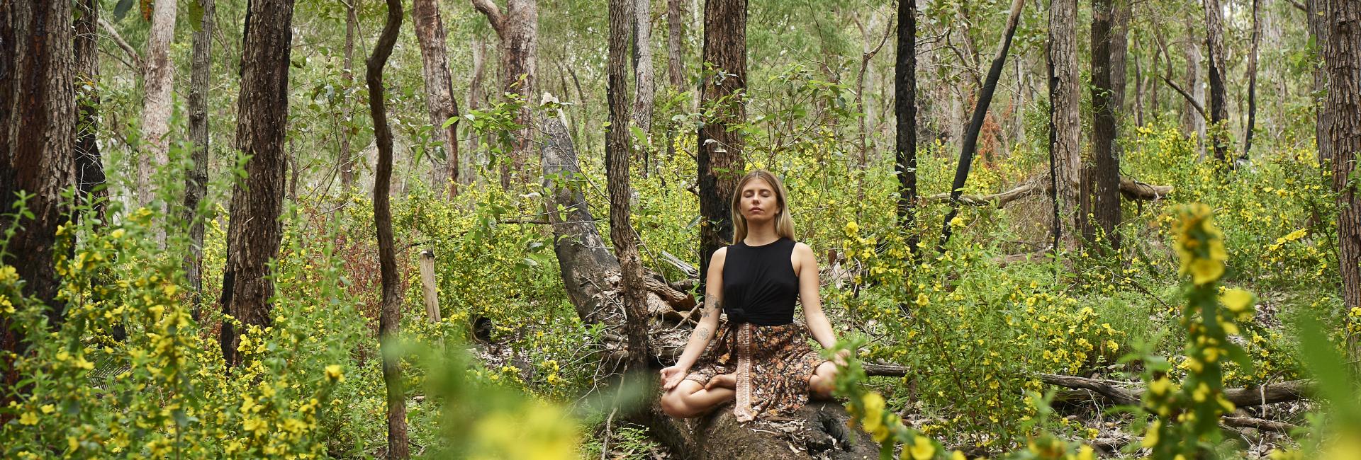 A girl sits cross legged meditating in a lush green forest