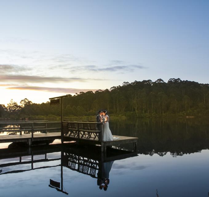 A bride and groom stand on a jetty with reflective water at sunset